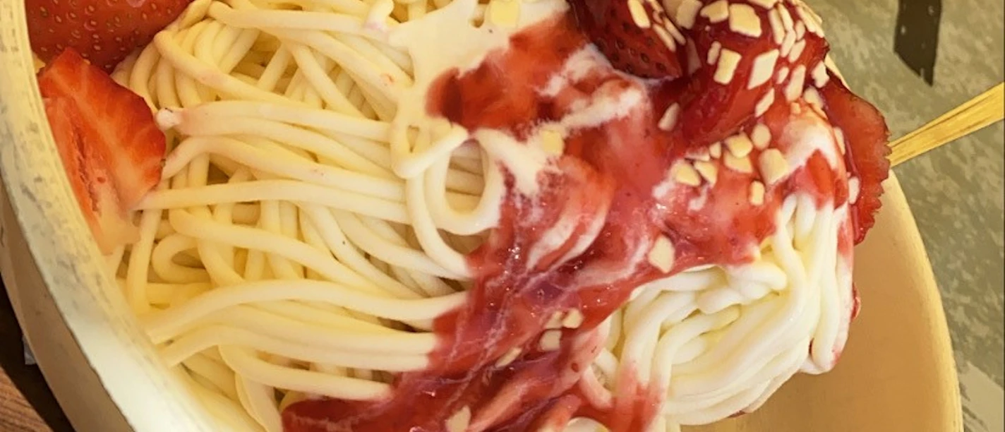 Eis Spaghetti-Eis mit Erdbeeren, Sahne, roter Erdbeersoße und Schokoraspeln in einer weißen Schale.