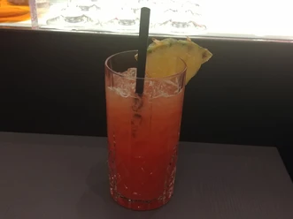 Ein bunt geschichteter Cocktail in einem Kristallglas, dekoriert mit Ananasstück und schwarzem Strohhalm.