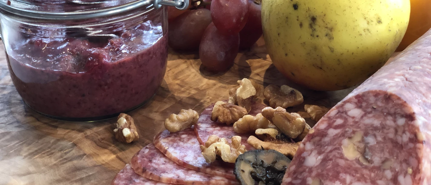 Kalte Platte Verschiedene Wurstsorten, Nüsse und Trauben liegen auf einem Holzbrett neben einem Glas Aufstrich.