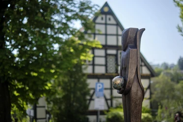 Hölzerne Skulptur einer Frau mit Kugel in der Hand, vor Fachwerkhaus und grünen Bäumen.
