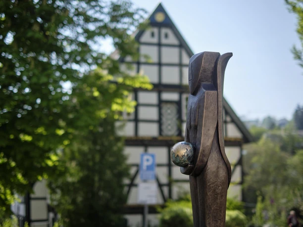 Hölzerne Skulptur einer Frau mit Kugel in der Hand, vor Fachwerkhaus und grünen Bäumen.