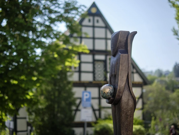 Die Wächterin Hölzerne Skulptur einer Frau mit Kugel in der Hand, vor Fachwerkhaus und grünen Bäumen.