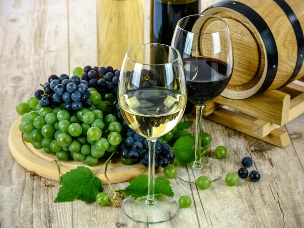 Zwei Weingläser, eines mit Weiß- und eines mit Rotwein, neben Trauben und einem Weinfass auf Holztisch.