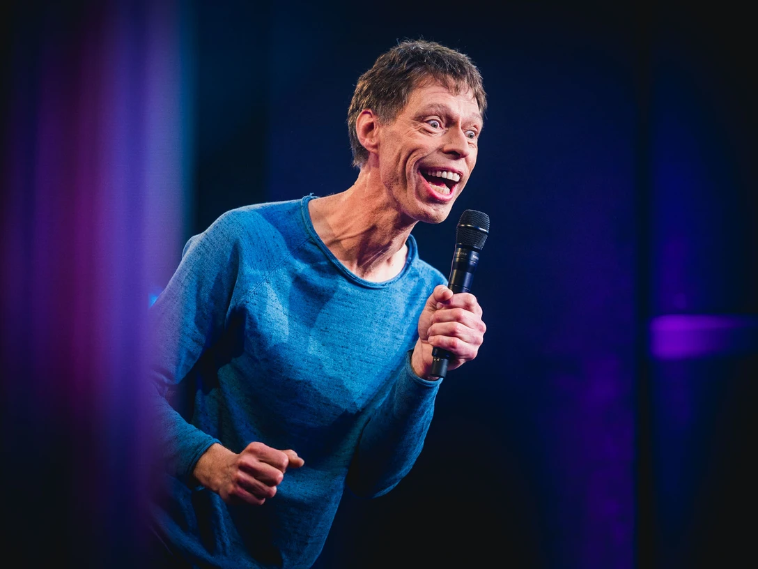 Maddin Schneider Kabarettist Maddin Schneider hält ein Mikrofon, lächelt und trägt ein blaues Sweatshirt.