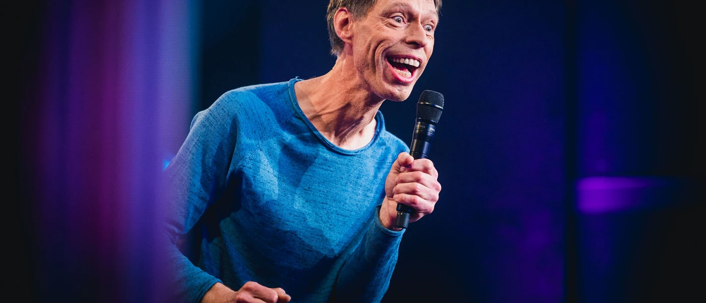 Maddin Schneider Kabarettist Maddin Schneider hält ein Mikrofon, lächelt und trägt ein blaues Sweatshirt.