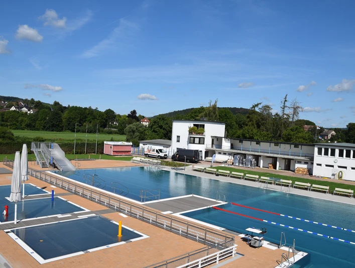 Freibad mit großem Schwimmbecken, Sprungturm und umliegenden Gebäuden, eingebettet in grüne Landschaft.