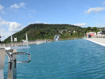 freibad_hoexter (2).JPG Freibad mit Blick auf bewaldeten Hügel, türkises Wasser, Sprungbrett und Rutsche bei Sonnenschein.