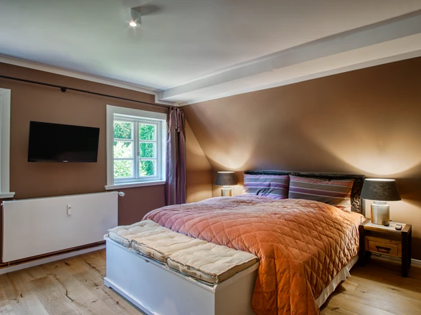 Schlafzimmer mit Doppelbett, oranger Tagesdecke, braunen Wänden, Fernseher und Holzboden.