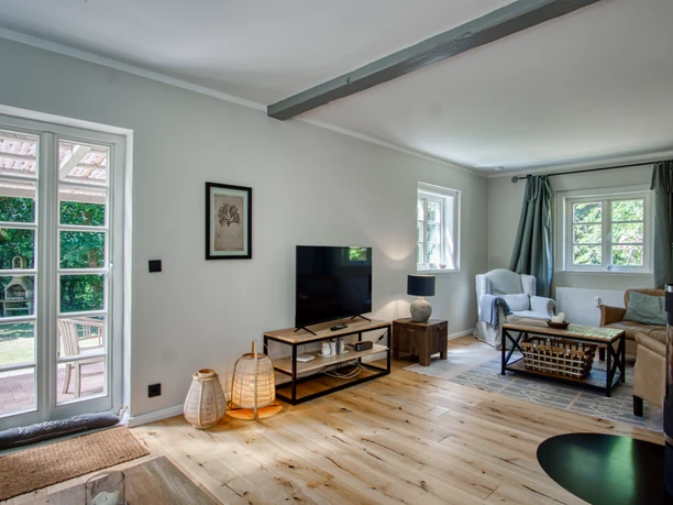 Heller Wohnbereich mit Holzfußboden, Fernseher, Sitzgruppe und einer Glastür zum grünen Garten.