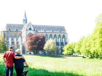Altenberger Dom Älteres Paar mit Rollator blickt auf eine gotische Kathedrale in einer grünen Landschaft.