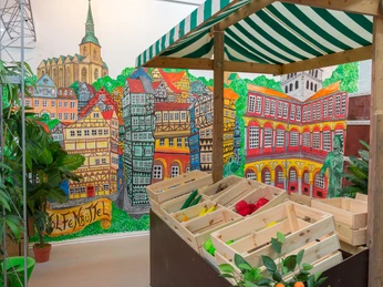 Aha_Museum-Ausstellung-3 Ein Marktstand. Die Wand ist bunt bemalt mit Fachwerkhäusern, dem Schloss und der HauptkircheA market stall. The wall is colorfully painted with half-timbered houses, the castle and the main churchEn markedsbod. Muren er farvestrålende malet med bindingsværkshuse, slottet og hovedkirken.Een marktkraam. De muur is kleurrijk beschilderd met vakwerkhuizen, het kasteel en de hoofdkerk