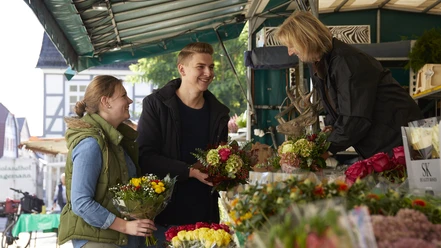 Wochenmarkt.jpg Menschen kaufen bunte Blumensträuße an einem belebten Marktstand unter einem Überdach.
