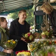 Wochenmarkt.jpg Menschen kaufen bunte Blumensträuße an einem belebten Marktstand unter einem Überdach.