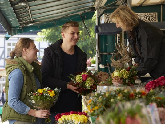Wochenmarkt.jpg Menschen kaufen bunte Blumensträuße an einem belebten Marktstand unter einem Überdach.