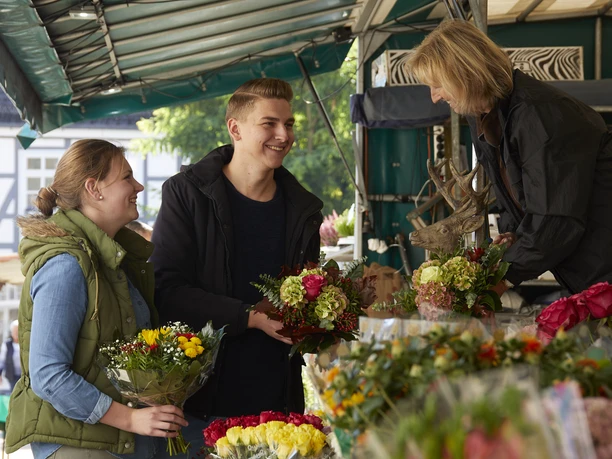 Wochenmarkt.jpg Menschen kaufen bunte Blumensträuße an einem belebten Marktstand unter einem Überdach.