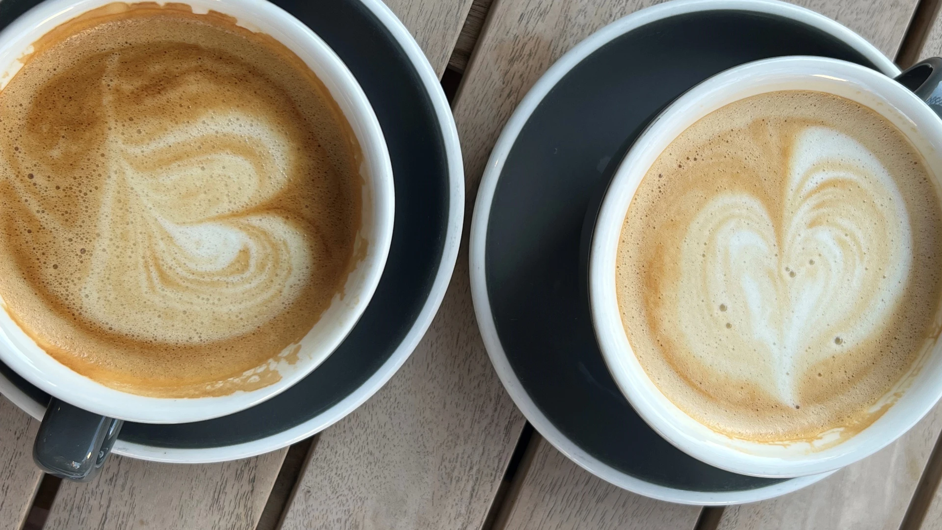 Zwei Kaffeetassen mit Latte Art in Herzform stehen auf grauen Tellern auf einem Holztisch.