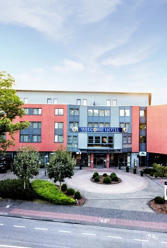 Das moderne Welcome Hotel Paderborn mit roter Fassade, großen Fenstern und grüner Umgebung.