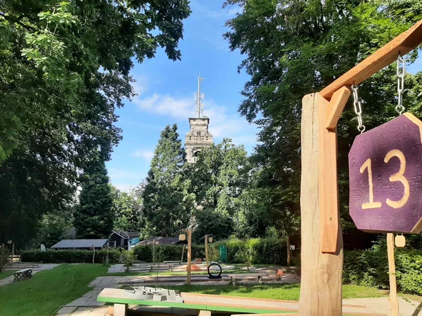 Minigolfplatz Waldkletterpark Velbert-Langenberg Ein Minigolfplatz mit Hindernis 13, umgeben von Bäumen, mit einem Turm im Hintergrund an einem sonnigen Tag.