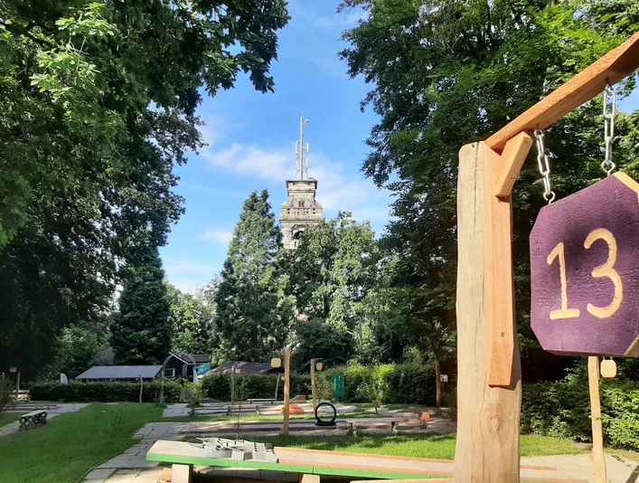 Minigolfplatz Waldkletterpark Velbert-Langenberg Ein Minigolfplatz mit Hindernis 13, umgeben von Bäumen, mit einem Turm im Hintergrund an einem sonnigen Tag.