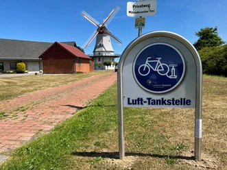 Lufttankstelle für Fahrräder vor der historischen Behlmer Mühle mit weißen Flügeln und rotem Dach.