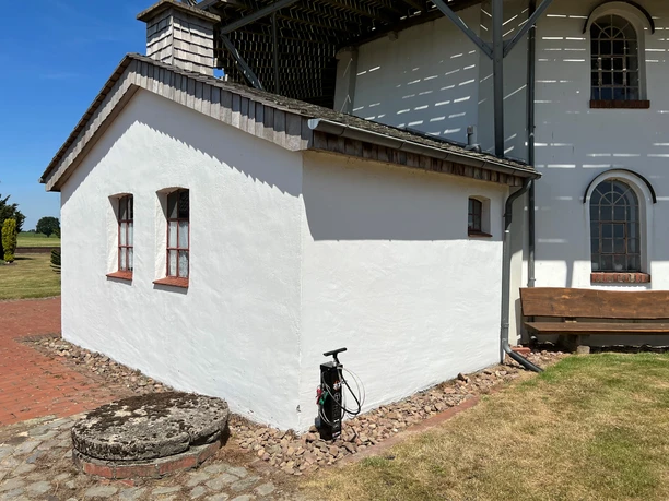 Lufttankstelle Behlmer Mühle Weiße Außenwand eines Gebäudes mit zwei Fenstern und einer Handluftpumpe auf gepflastertem Boden.