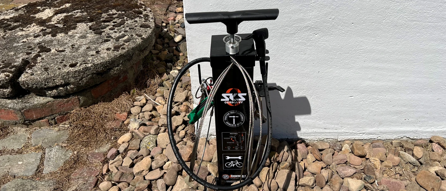 Lufttankstelle Behlmer Mühle Fahrradpumpe neben einer weißen Wand, umgeben von Pflastersteinen und im Hintergrund liegt eine gepflasterte Fläche.