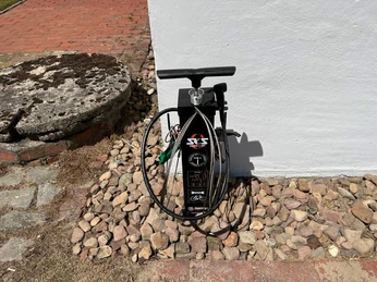 Lufttankstelle Behlmer Mühle Fahrradpumpe neben einer weißen Wand, umgeben von Pflastersteinen und im Hintergrund liegt eine gepflasterte Fläche.