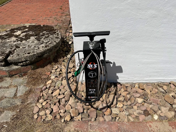 Lufttankstelle Behlmer Mühle Fahrradpumpe neben einer weißen Wand, umgeben von Pflastersteinen und im Hintergrund liegt eine gepflasterte Fläche.