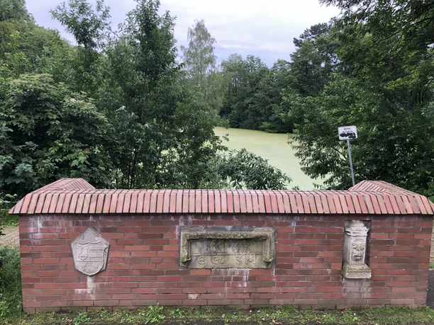 Sedanbrücke Backsteinmauer am Teichweg. Reliefs und ein Schild dekorieren die Mauer mit Blick auf den Teich.