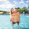 Freibad Angerbad in Ratingen Zwei Personen in einem Außenpool lächeln fröhlich, umgeben von Bäumen und einem klaren, blauen Himmel.