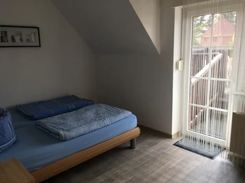 Ferienwohnung am Schafstall Dachschräge mit Einzelbett, blauer Bettwäsche, kleinem Balkon und großem Fenster mit Vorhang.