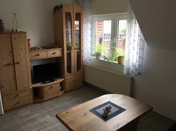 Gemütliches Wohnzimmer mit Holzmöbeln, Fenster mit Pflanzen und einem kleinen Fernseher auf Kommode.