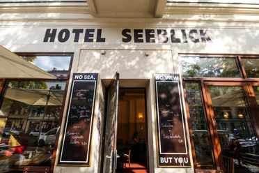 Hotel Seeblick - Gastronomie in Leipzig Blick auf den Außenbereich des Hotel Seeblicks, den Gehweg, die Häuserfassade und mehr mit Sonnenschirmen, Stühlen und Co. an einem Sommertag; mit Blick auf den Straßenzug und angrenzende Häuser, Gastronomie, kulinarik, restaurant, freisitz
