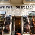 Hotel Seeblick - Gastronomie in Leipzig Blick auf den Außenbereich des Hotel Seeblicks, den Gehweg, die Häuserfassade und mehr mit Sonnenschirmen, Stühlen und Co. an einem Sommertag; mit Blick auf den Straßenzug und angrenzende Häuser, Gastronomie, kulinarik, restaurant, freisitz