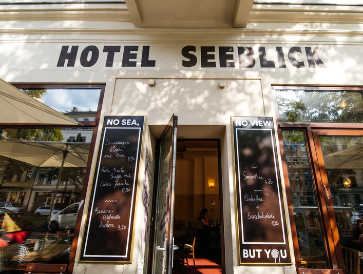 Hotel Seeblick - Gastronomie in Leipzig Blick auf den Außenbereich des Hotel Seeblicks, den Gehweg, die Häuserfassade und mehr mit Sonnenschirmen, Stühlen und Co. an einem Sommertag; mit Blick auf den Straßenzug und angrenzende Häuser, Gastronomie, kulinarik, restaurant, freisitz