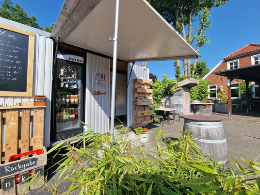 Pausenstopp in Marienheil, Rhauderfehn, Ostfriesland Kleiner Außenbereich eines Pausen-Cafés mit Getränkekühlschrank, Holztisch, Fass als Stehtisch und Pflanzen unter blauem Himmel.
