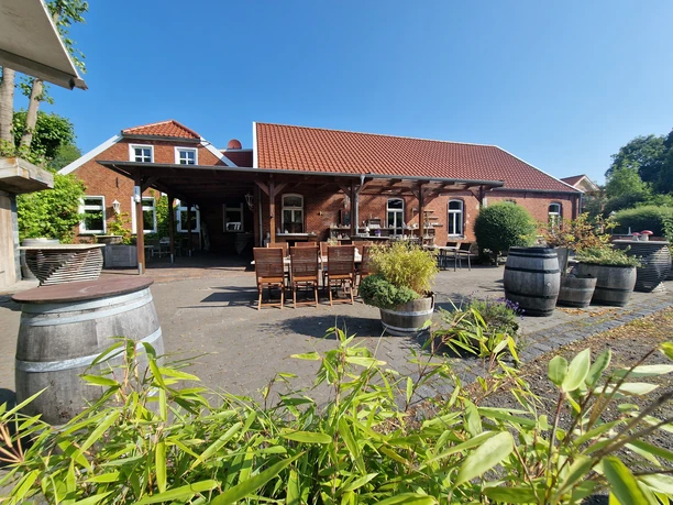 Pausenstopp in Marienheil, Rhauderfehn, Ostfriesland Außenbereich eines Gasthauses mit roten Ziegeldächern, Holztischen und Stühlen auf einer Terrasse, umgeben von großen Pflanzgefäßen und grünem Bewuchs unter klarem blauem Himmel.