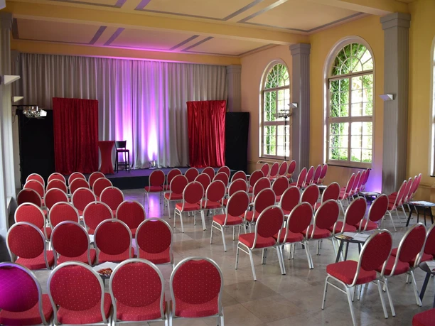 Bad Driburg_Theater im Park_Bühne in den Brunnenarkaden Roter Theatersaal mit Parkettboden, gefüllten Stuhlreihen und Bühne in elegantem Saal mit hohen Fenstern.