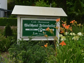 Ein hölzernes Schild mit der Aufschrift "Waldhotel Felsenkeller" neben blühenden orangefarbenen Lilien.
