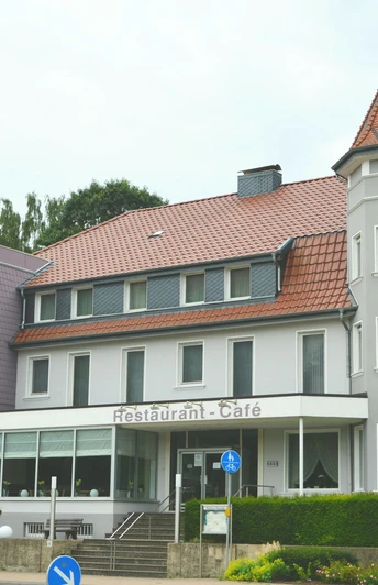 Außenansicht eines mehrstöckigen Hotels mit Türmchen, Waldbewuchs im Hintergrund, Fassade in Weiß-Grau.