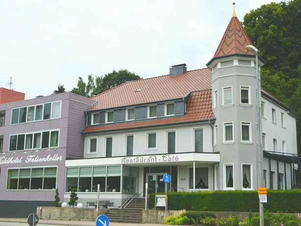 Außenansicht eines mehrstöckigen Hotels mit Türmchen, Waldbewuchs im Hintergrund, Fassade in Weiß-Grau.