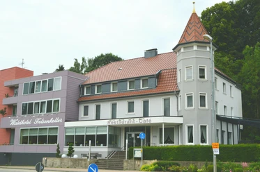 Hotel Felsenkeller_Bad Iburg_Teutoburger Wald Tourismus_I Bohlken (3)_3000.jpg Außenansicht eines mehrstöckigen Hotels mit Türmchen, Waldbewuchs im Hintergrund, Fassade in Weiß-Grau.