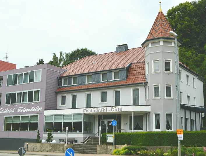 Hotel Felsenkeller_Bad Iburg_Teutoburger Wald Tourismus_I Bohlken (3)_3000.jpg Außenansicht eines mehrstöckigen Hotels mit Türmchen, Waldbewuchs im Hintergrund, Fassade in Weiß-Grau.