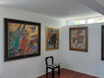 Museum Raffael Becker Das Bild zeigt die Innenansicht eines Ausstellungsraums im Museum Raffael Becker. An den strahlend weißen Wänden sind verschiedene, farbenfrohe Gemälde mit dicken Rahmen angebracht. Der Boden besteht aus dunklem Holz. Ein einzelner schwarzer Stuhl mit gestreifter Polsterung steht in der Ecke des Raumes, unter den Gemälden, als ob er die Kunstwerke zum Betrachten einlädt.The picture shows the interior of an exhibition room in the Raffael Becker Museum. Various colorful paintings with thick frames are mounted on the bright white walls. The floor is made of dark wood. A single black chair with striped upholstery stands in the corner of the room, beneath the paintings, as if inviting the works of art to be viewed.