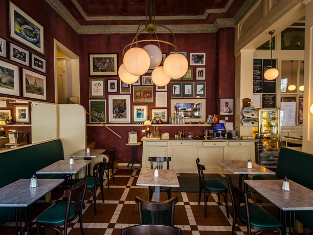 Café Maître - Kawiarnie w Lipsku Widok na historyczną jadalnię Café Maître z meblami z kutego żelaza w stylu secesyjnym i wieloma zdjęciami na ciemnoczerwonych ścianach, kawiarnia, gastronomia, restauracja