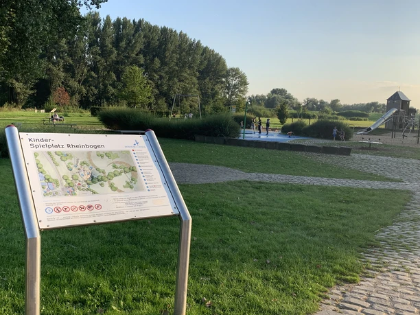 Rheinbogen mit Wasserspielplatz in Monheim am Rhein Freizeitfläche mit Spielplatz in grüner Umgebung am Rhein, Spielgeräte und Besucher im Hintergrund.