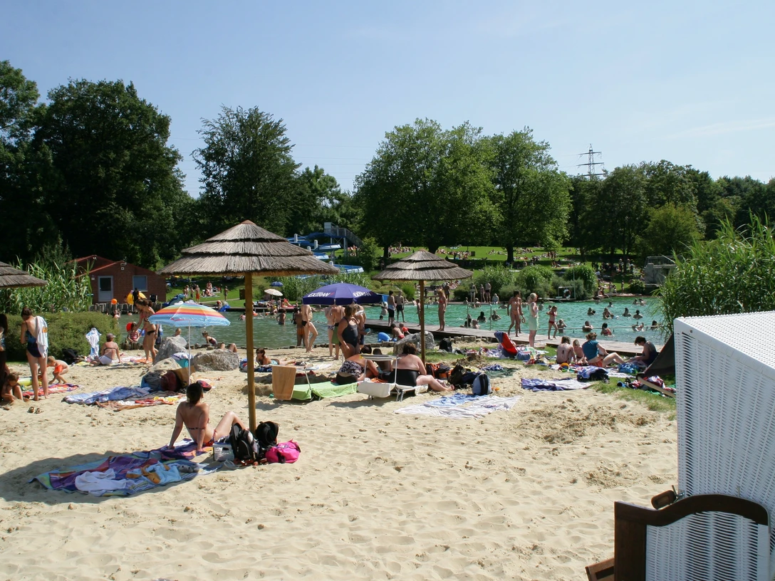 Naturfreibad Mettmann Ein Naturfreibad mit Sandstrand und Sonnenschirmen, umgeben von Bäumen und entspannenden Gästen.