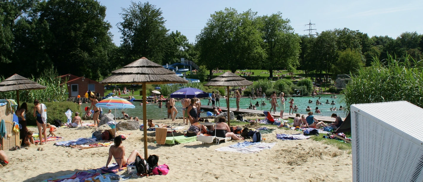 Naturfreibad Mettmann Ein Naturfreibad mit Sandstrand und Sonnenschirmen, umgeben von Bäumen und entspannenden Gästen.