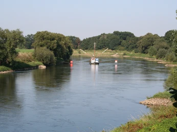 Drakenburg Blick über einen ruhigen Fluss mit einem angedockten Boot umgeben von grüner Landschaft und Bäumen.