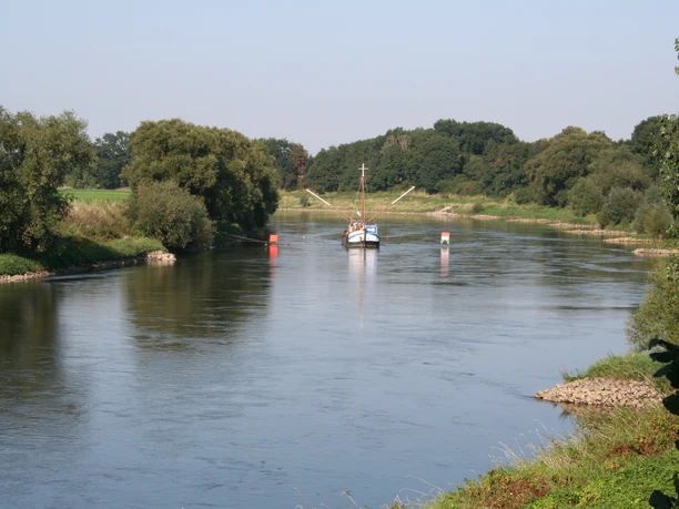 Drakenburg Blick über einen ruhigen Fluss mit einem angedockten Boot umgeben von grüner Landschaft und Bäumen.